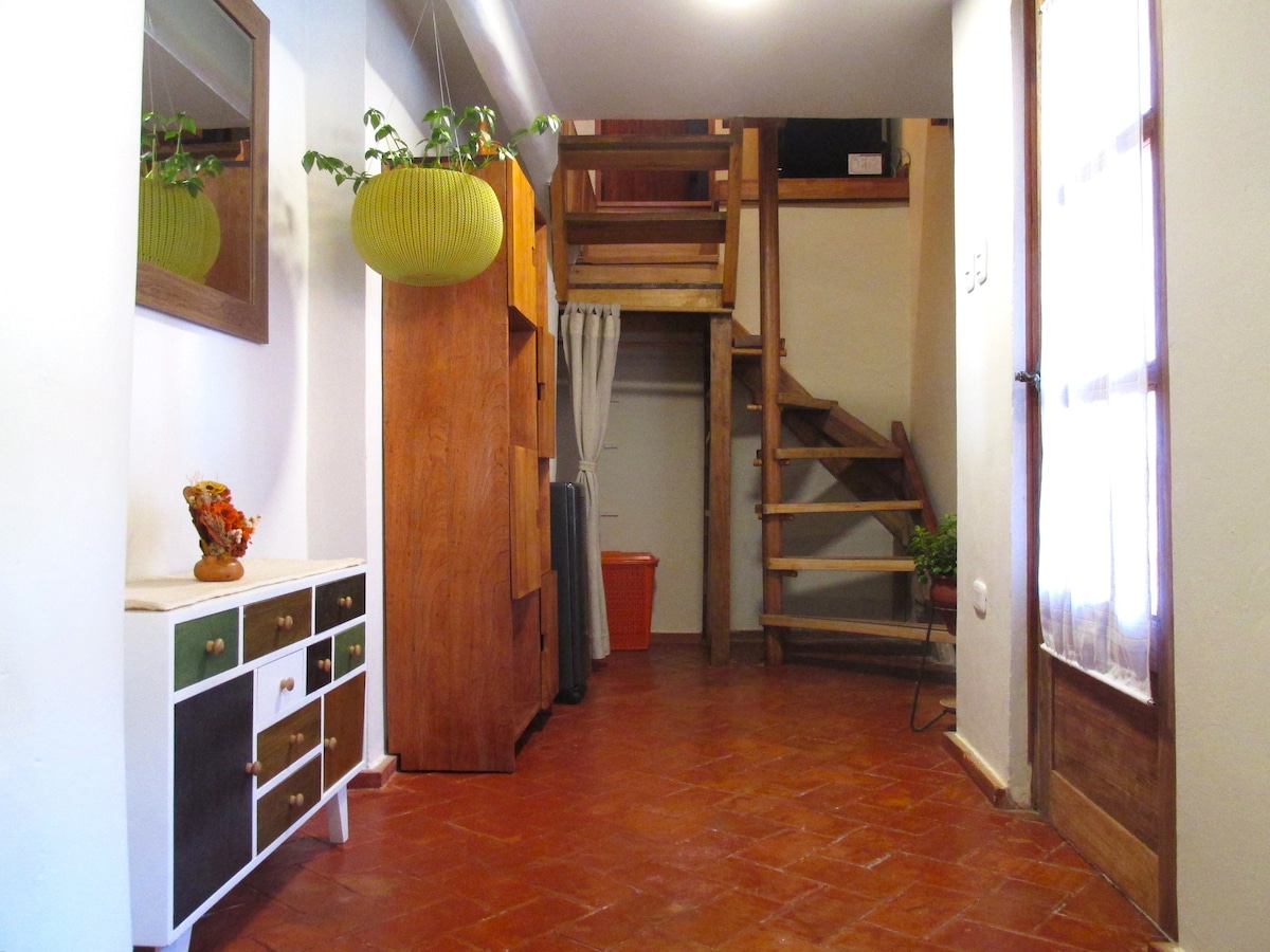 The entrance area features a warm-toned tiled floor and simple wooden furnishings. A console table with colorful drawers is positioned against the wall, while hanging plants add a touch of greenery. A staircase leads to the upper level, inviting guests to explore further.