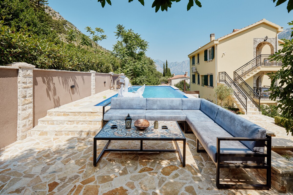 An inviting outdoor area features a modern lounge set with a low table, surrounded by stone paving. A private pool is positioned in the background, framed by lush greenery and mountain views, with a stone wall providing a sense of seclusion.