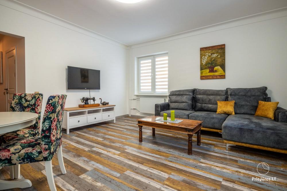 The spacious living area features a comfortable gray sofa and a wooden coffee table. A flat-screen TV is mounted on the wall opposite. Floral-patterned chairs are positioned around a round dining table. Natural light enters through a window with horizontal blinds.