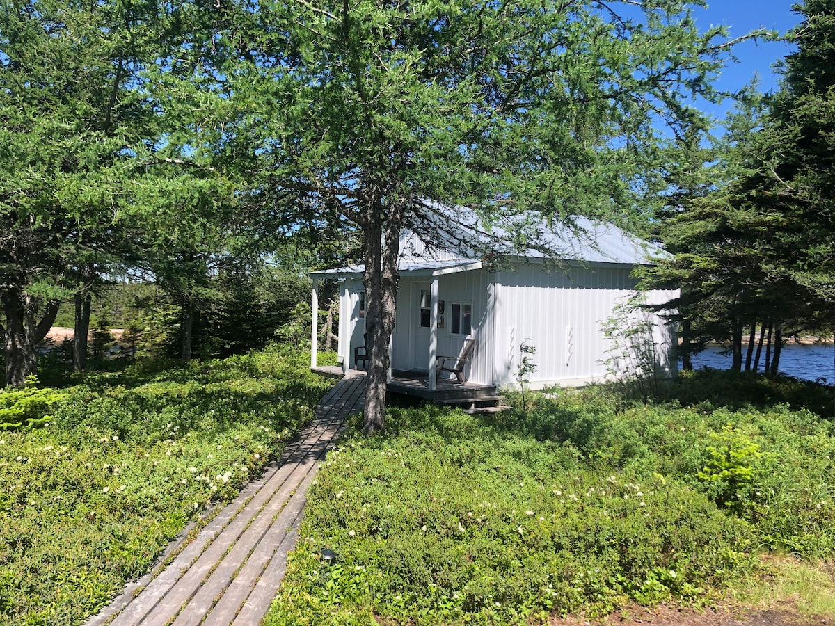 A private chalet is nestled among lush greenery, featuring a light-colored exterior with a metal roof. A wooden pathway leads to the entrance, which is flanked by Adirondack chairs. The surrounding landscape includes trees and a view of the water nearby.