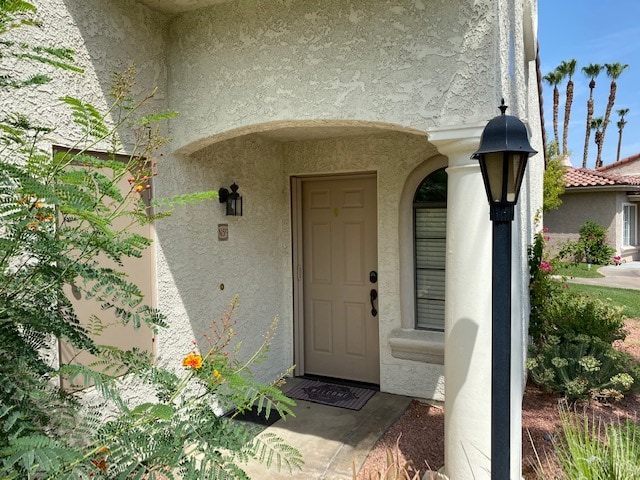 The entrance to the condo is framed by a shaded porch area, featuring an arched entryway and a welcoming door. A soft light is provided by a lamp post beside the path, with lush greenery and blooming plants enhancing the serene atmosphere.