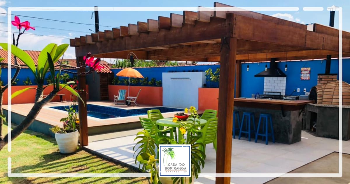 An outdoor space is presented with a wooden pergola overseeing a swimming pool. Seating for six is available at a green dining set, complemented by an orange parasol. A barbecue area is visible, featuring a grill and stone surface, surrounded by colorful plants.