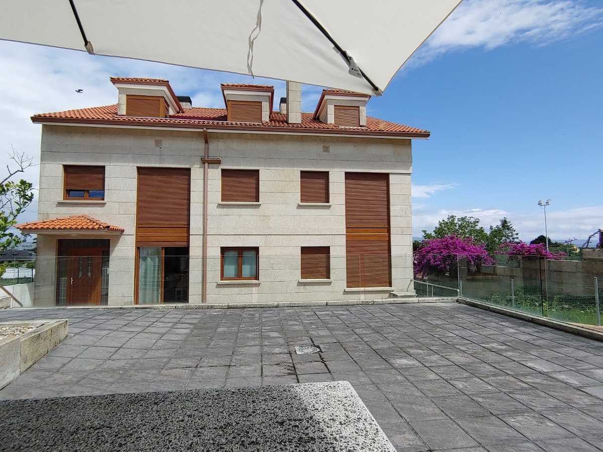The exterior of a two-story stone house is displayed, featuring a warm-toned façade with wooden shutters. A paved terrace surrounds the structure, providing ample outdoor space. Bright bougainvillea is visible nearby, with a clear blue sky above and a few clouds.
