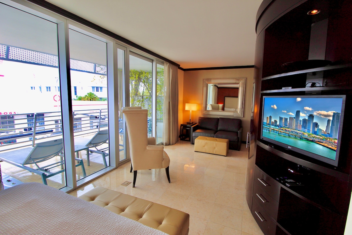 An inviting suite features large glass doors leading to a private balcony, allowing natural light to fill the space. A comfortable seating area with a dark sofa and elegant chair complements the sleek design. A TV is mounted on the wall, enhancing the entertainment options.