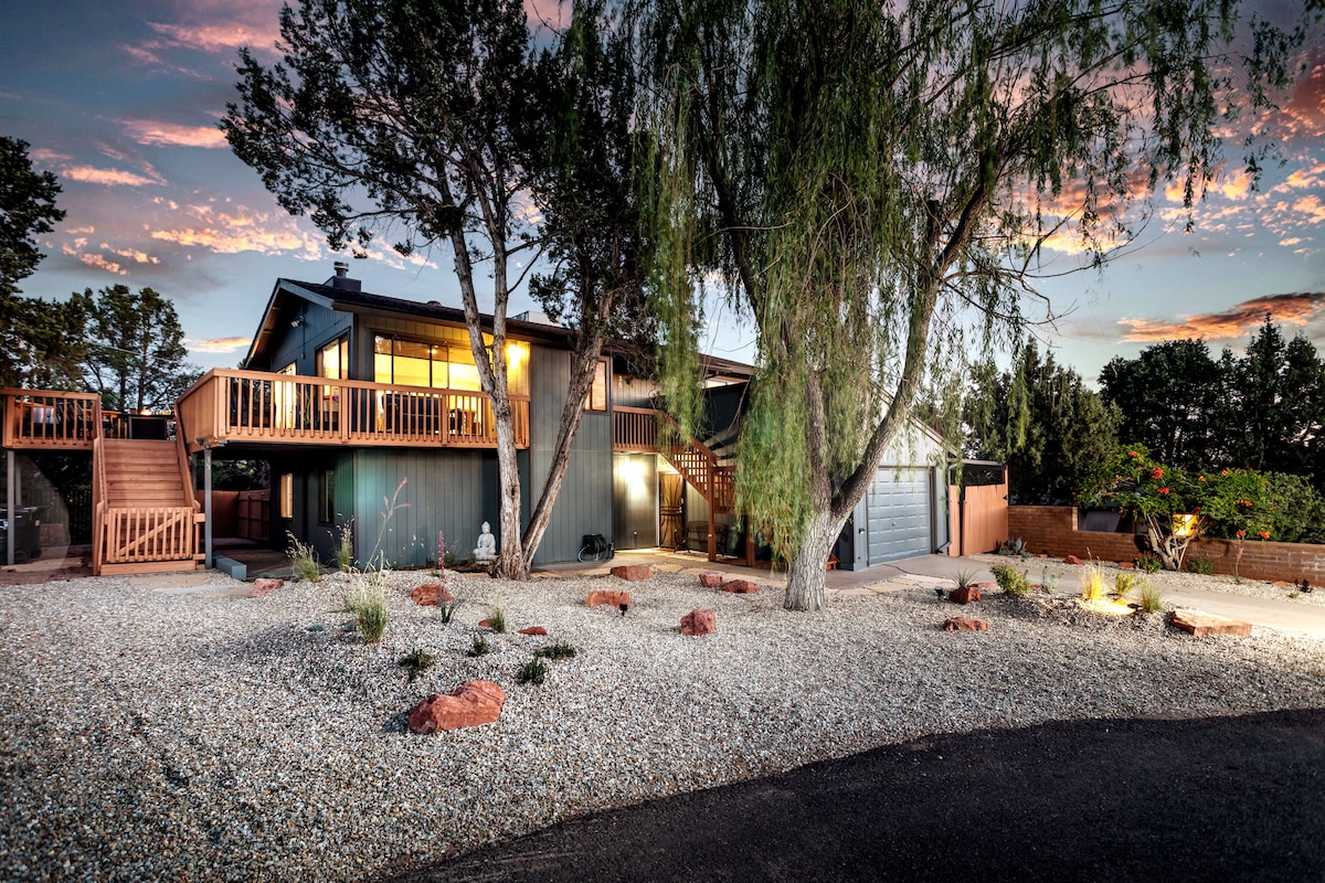 A two-story home is depicted, featuring a broad staircase leading to an upper deck that is accessible from the exterior. Surrounding landscaping includes gravel, scattered red rocks, and various plants. The structure's exterior is accented by a strong, warm sunset sky.