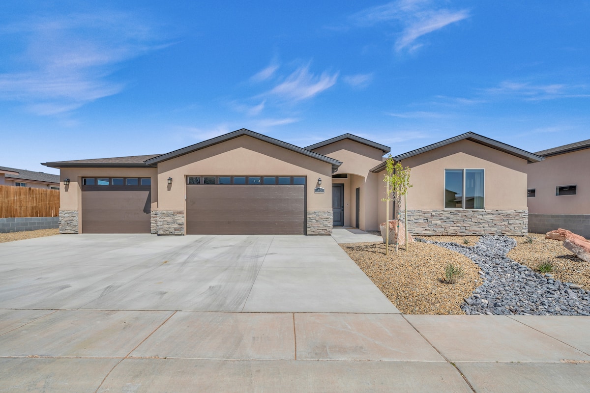 A modern single-story home features a broad facade with two garage doors and a welcoming front entrance. The exterior is complemented by stone accents, desert landscaping with gravel and minimal vegetation, and a clear blue sky in the background.