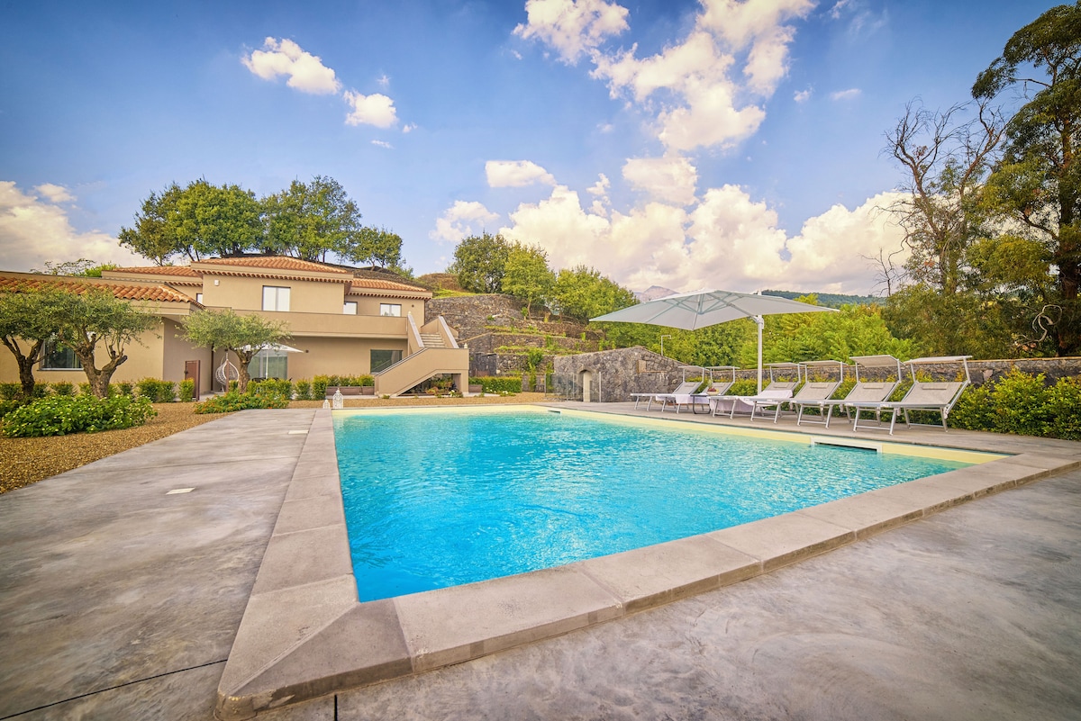 A rectangular salt-water pool is surrounded by a spacious deck with several lounge chairs. Lush greenery and stone features create a natural setting, while a smooth pathway leads to a nearby building. The sky is filled with clouds, enhancing the serene atmosphere.