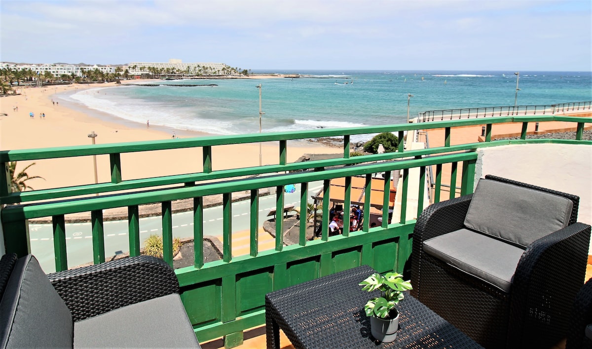 A balcony view showcases a sandy beach and turquoise waters, framed by a green railing. Comfortable outdoor furniture is positioned for relaxation, with a small potted plant adding a touch of greenery. The vibrant coastal scene conveys a serene atmosphere.