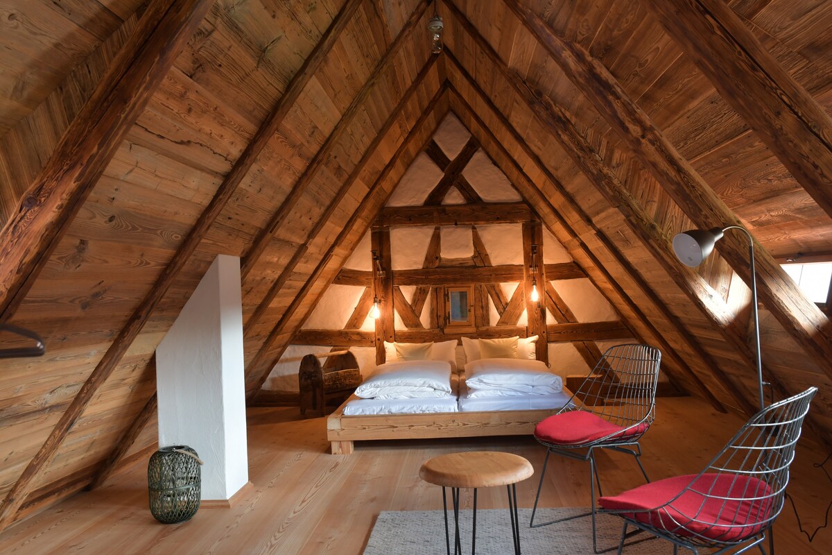 An airy loft bedroom features a high, sloping wooden ceiling and exposed beams. A spacious bed with white linens is positioned centrally, flanked by two modern chairs. Soft lighting from wall-mounted fixtures creates a relaxed ambiance, while a small round table sits nearby on a textured area rug.