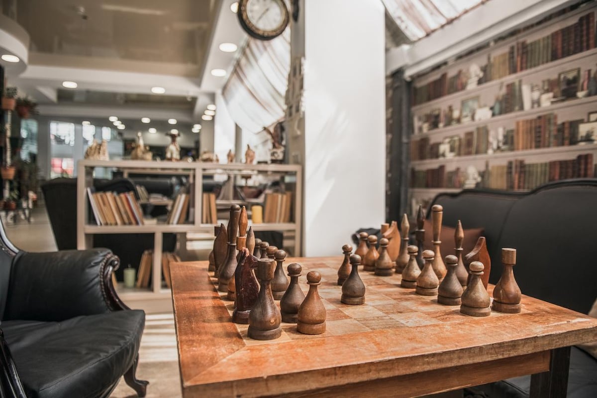 A wooden chess table is positioned prominently in a cozy sitting area. The intricately carved chess pieces sit in formation, ready for play. Bookshelves line the background, filled with a collection of books, enhancing the relaxed environment.