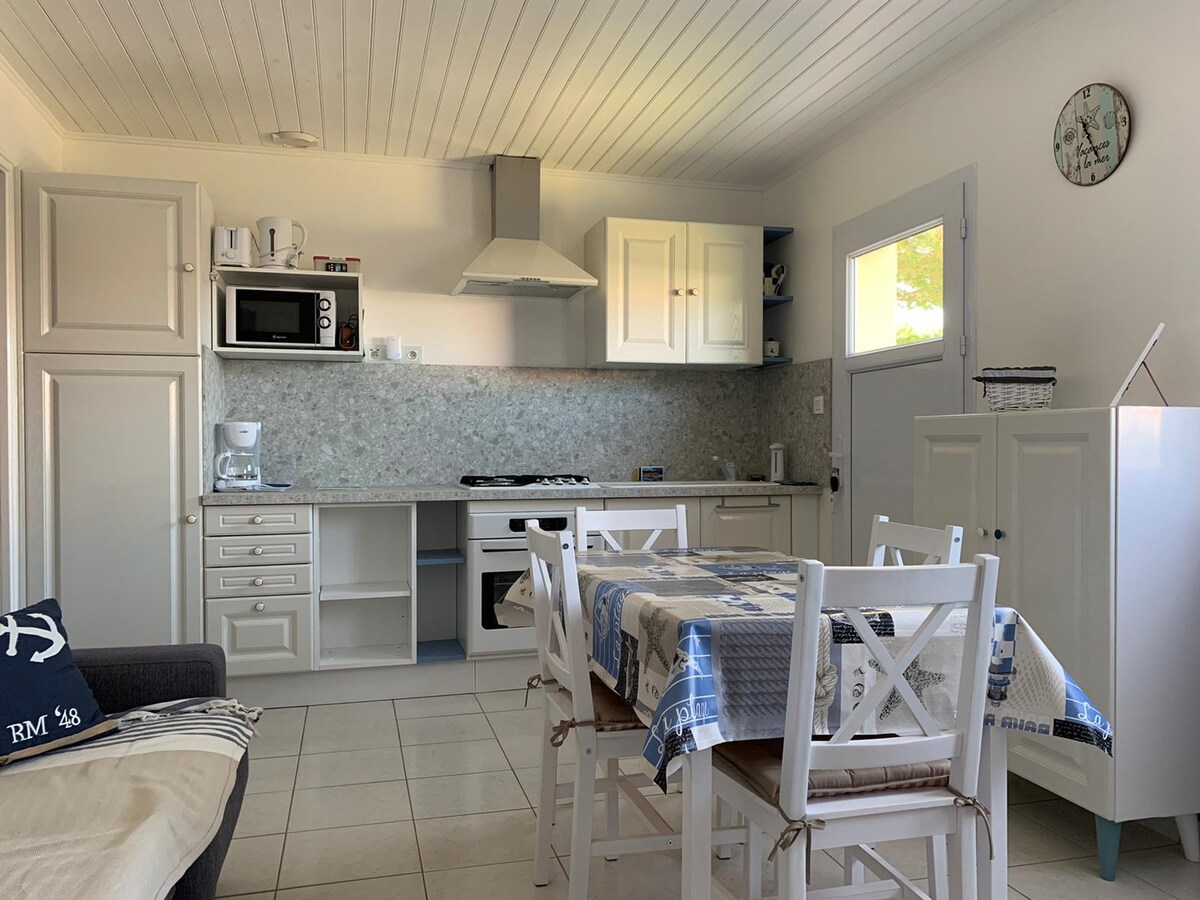 A well-equipped kitchen is shown, featuring a modern gas stove, microwave, and refrigerator. White cabinetry complements a light-colored countertop. A dining table surrounded by white chairs is positioned nearby, with natural light filtering through a window, enhancing the room's functionality.