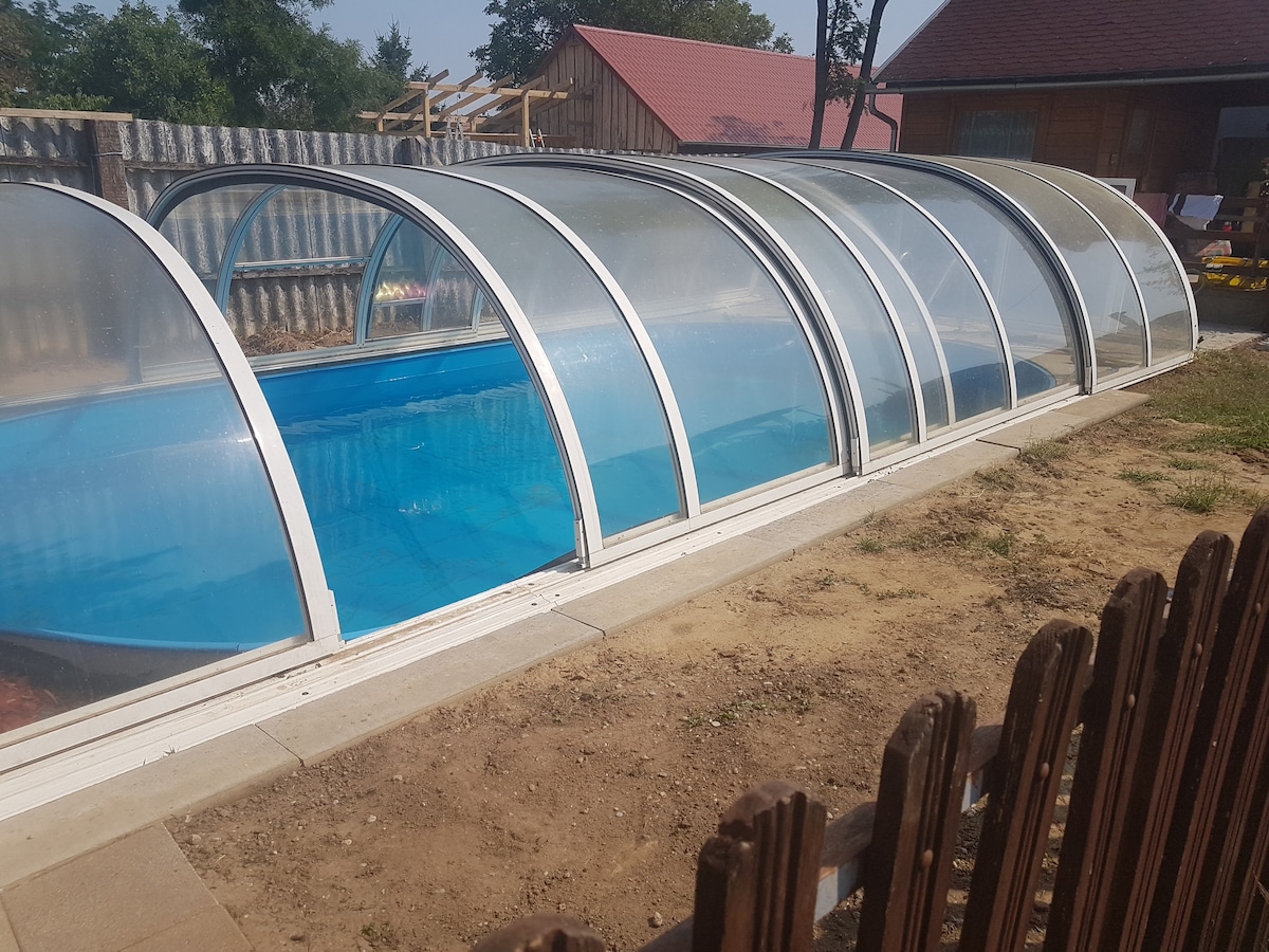 A covered swimming pool is displayed, featuring a transparent dome structure that allows natural light to illuminate the water. The surrounding area is well-kept, with a smooth paved edge and grassy patches, contributing to a serene outdoor setting.