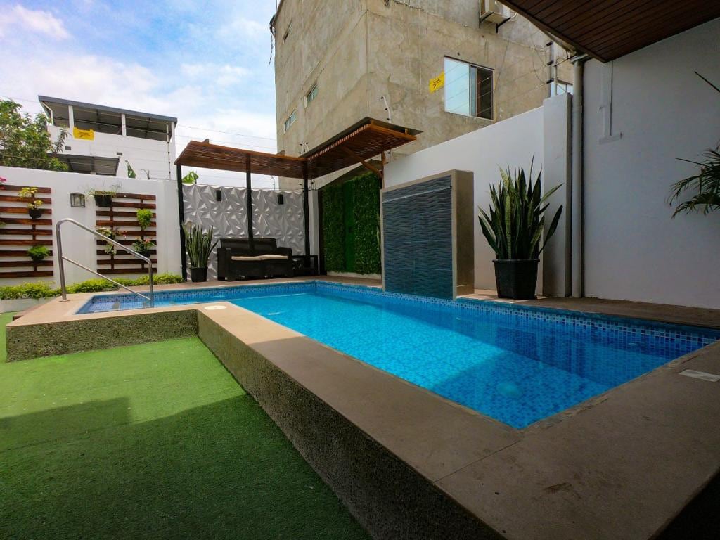 A private pool is framed by soft green grass and tall plants, offering a refreshing blue water contrast. A seating area with dark cushions is positioned under a wooden pergola, providing shaded relaxation beside the water feature.