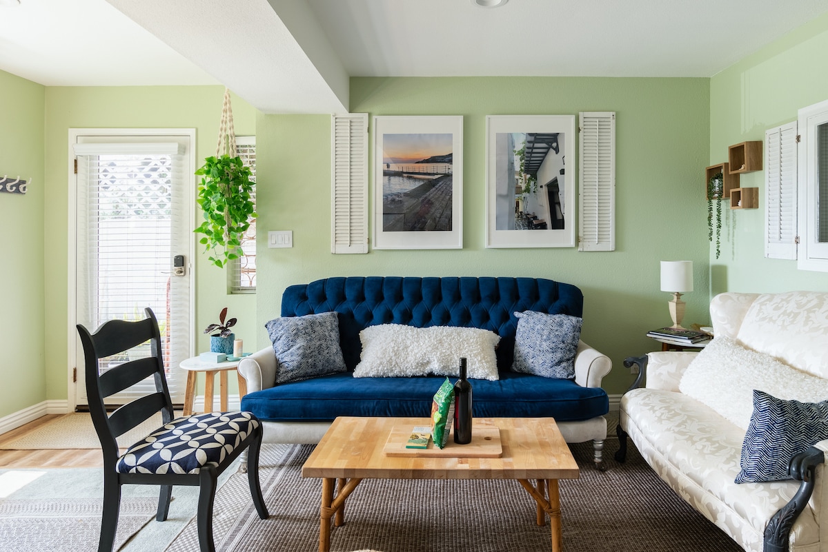 The living area features a blue velvet sofa and a white upholstered couch, positioned around a wooden coffee table. Two framed photographs adorn the green walls, with light filtering through nearby windows. A decorative plant hangs in one corner, adding a touch of greenery.