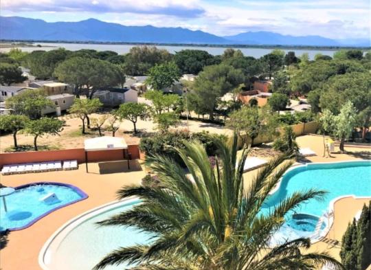 A panoramic view showcases the outdoor swimming pool area surrounded by lush trees and well-maintained gardens. Blue waters contrast with landscaped pathways, leading to units and the distant backdrop of mountains and a serene body of water.