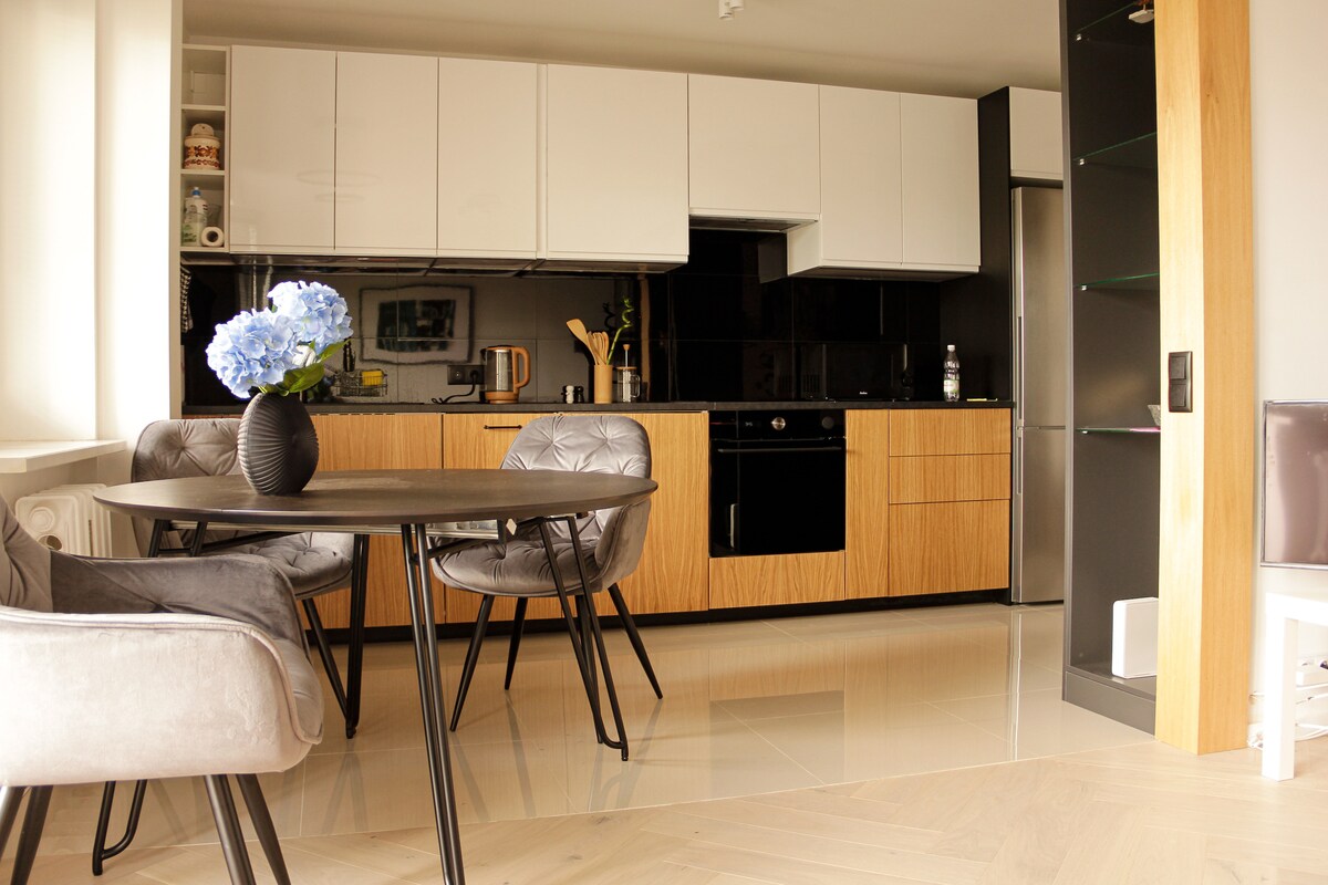 A modern kitchen area is featured, showcasing a sleek black countertop and light wood cabinetry. A round dining table with four chairs is placed nearby, complemented by a vase of blue flowers. Large windows offer natural light, enhancing the spacious feel.