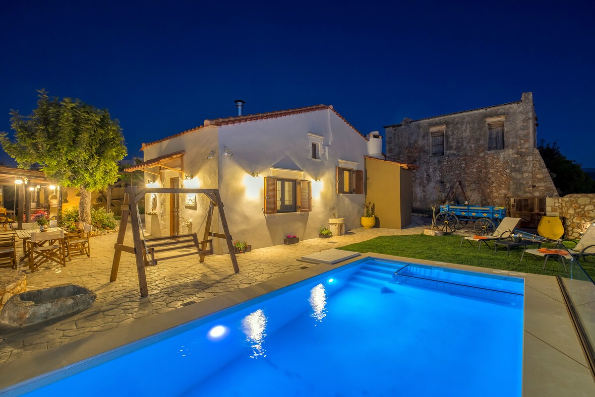 A traditional stone cottage is highlighted against a night sky, with soft lighting illuminating the exterior. A pool reflects the blue water under the stars. A swing set and seating area are visible in the yard, enhancing the outdoor space.