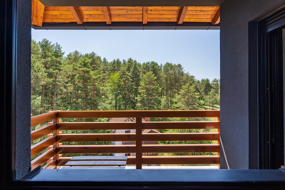A wooden balcony is framed by an opening, offering a view of lush green trees in the distance. The natural scenery provides a serene backdrop, emphasizing the peaceful surroundings of the apartment. Bright daylight enhances the inviting ambiance of the outdoor space.