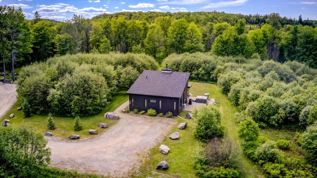 An aerial view captures a secluded black chalet surrounded by lush greenery. A gravel pathway leads to the building, bordered by natural stones. The expansive landscape showcases a mix of trees and shrubbery, providing a sense of privacy and tranquility in a serene environment.