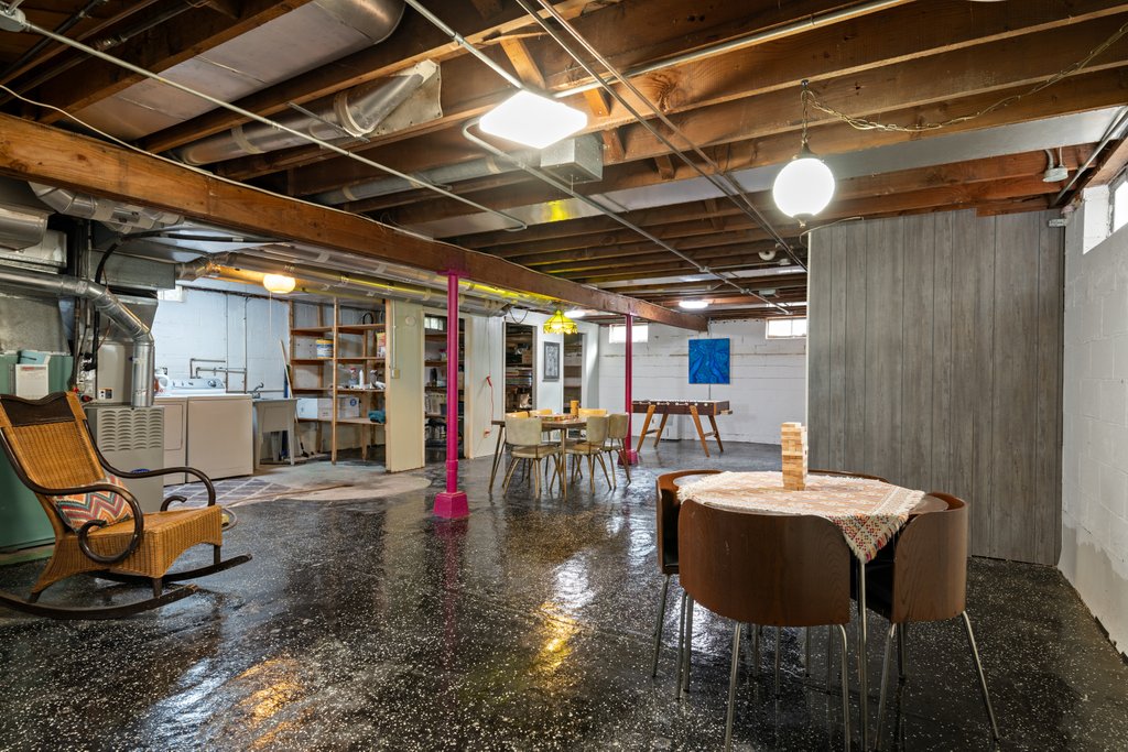 The spacious basement features a polished floor and a mix of functional areas. A dining table with chairs is adjacent to a cozy rocking chair. Shelving units provide storage, while laundry appliances are visible in the background. Bright lighting creates an inviting atmosphere.