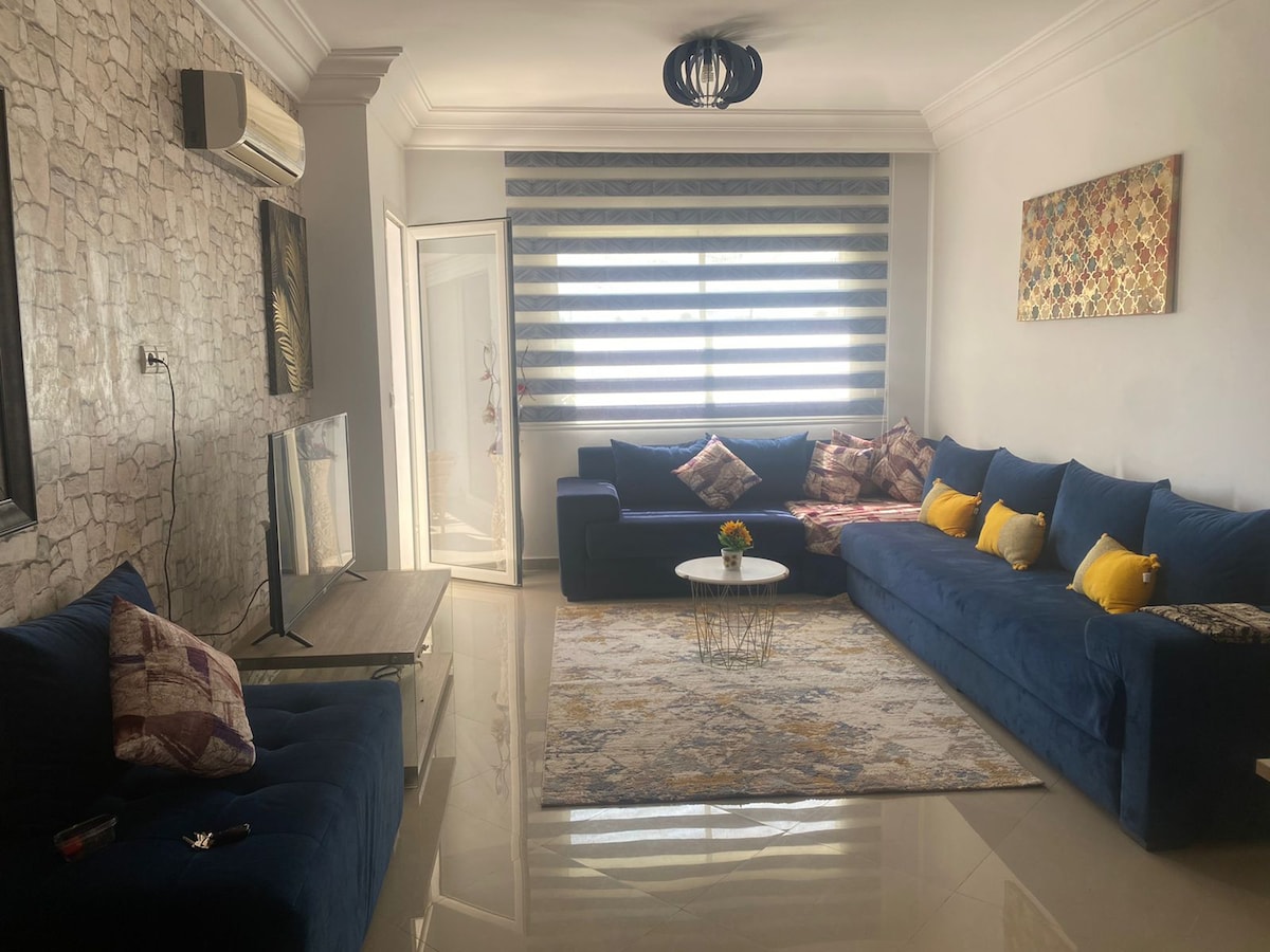 A spacious living room features a large sectional sofa adorned with colorful cushions. A coffee table is positioned on a decorative rug. Natural light filters through the window treatments, illuminating the room. A television is mounted on the wall, and artwork decorates the space.