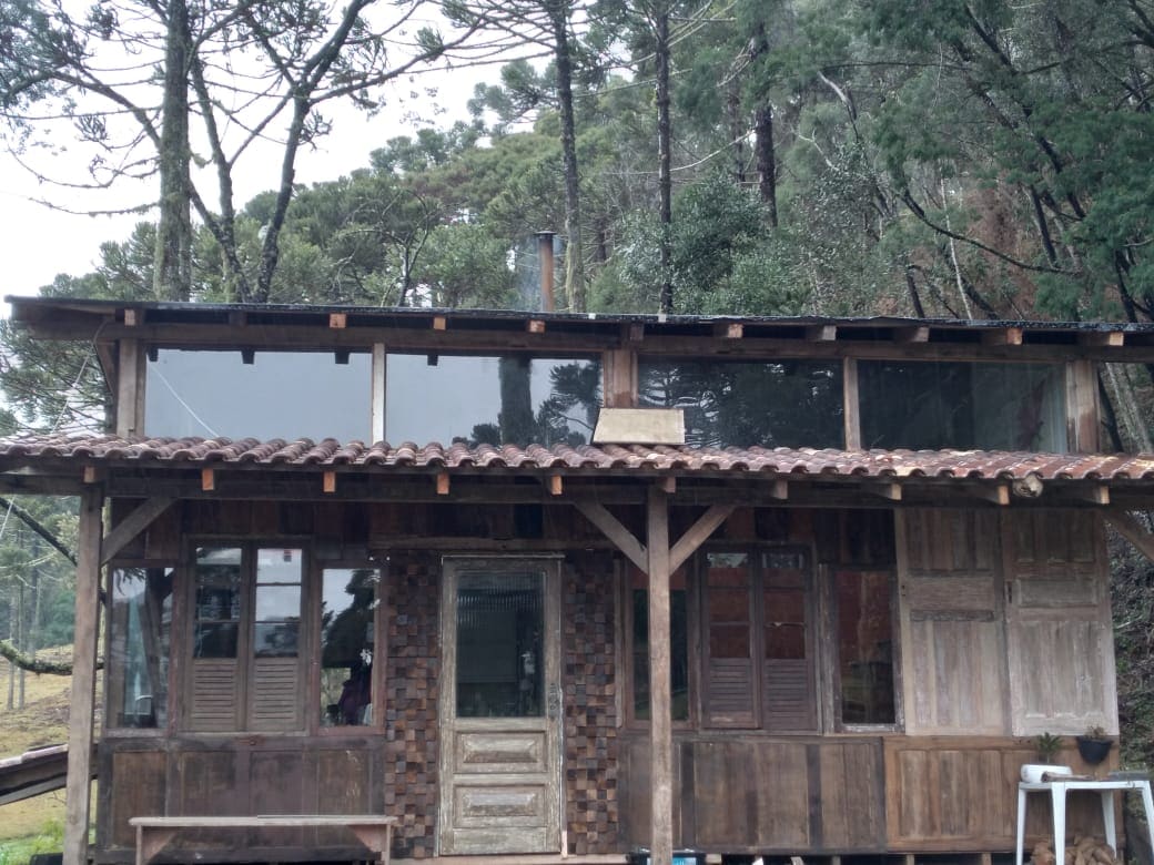 A rustic wooden cabin is presented, featuring large windows that allow natural light to illuminate the interior. The structure is supported by a wooden porch, with a tiled roof above. Surrounding the cabin, tall trees provide a natural backdrop.
