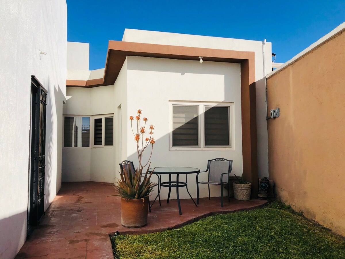 An outdoor patio is depicted, featuring a small table with two chairs. Surrounding greenery includes potted plants, contrasting with the terracotta flooring. A beige wall provides privacy, while a clear blue sky enhances the bright ambiance.