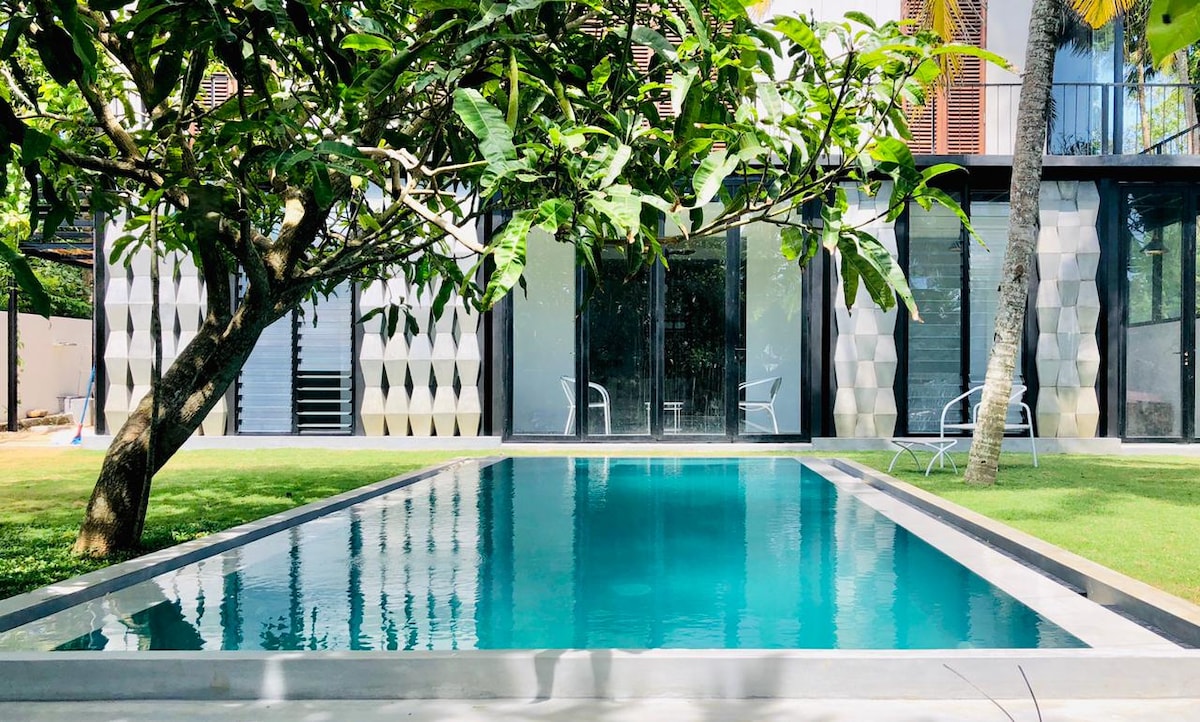 A serene garden pool is framed by lush greenery and palm trees, reflecting the clear blue sky. A modern building with large glass doors is visible in the background, creating a light-filled atmosphere. Comfortable seating is positioned nearby on the softly manicured lawn.