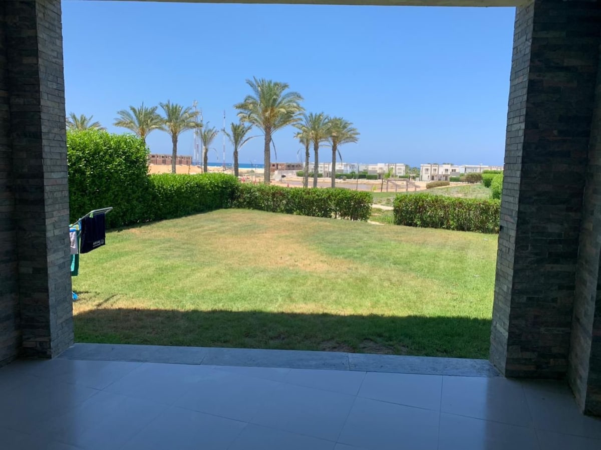 An outdoor view is presented from a covered patio, showcasing a well-maintained lawn bordered by lush green hedges. In the distance, palm trees and a glimpse of the ocean can be seen under a clear blue sky.