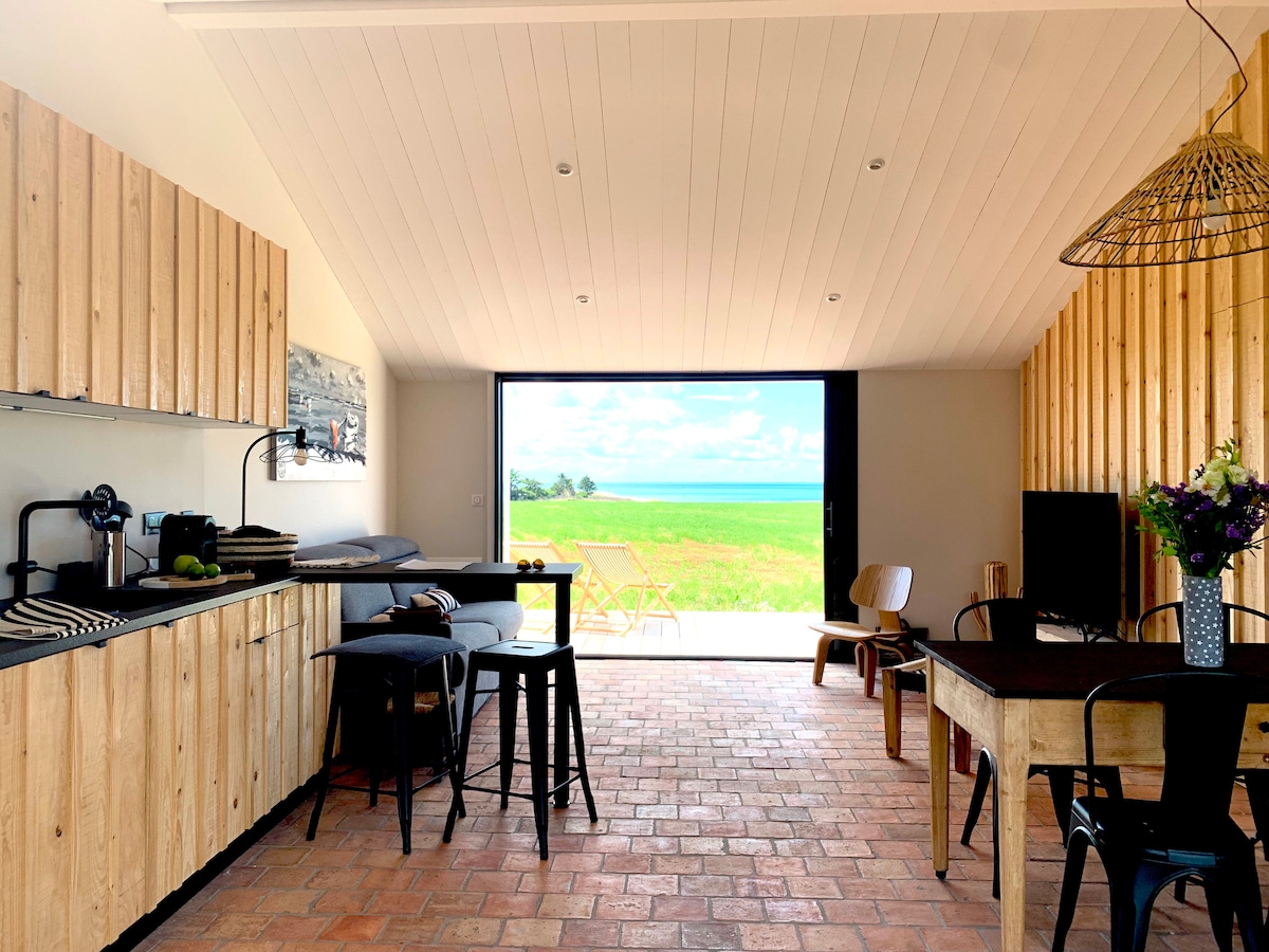 An inviting open-plan living area features wooden cabinetry and ample natural light. A dining table and chairs are positioned alongside a cozy seating area. Sliding glass doors lead to a picturesque outdoor view of the ocean and surrounding greenery, enhancing the space's connection to nature.