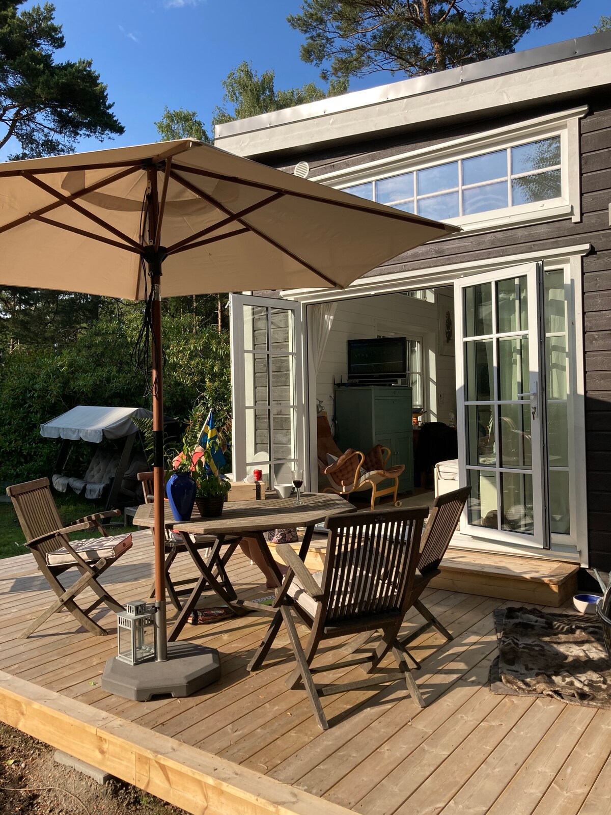 A wooden deck is furnished with a dark wooden table and four matching chairs, all shaded by a large umbrella. Behind the seating area, large glass doors provide a glimpse into the interior, surrounded by lush greenery.