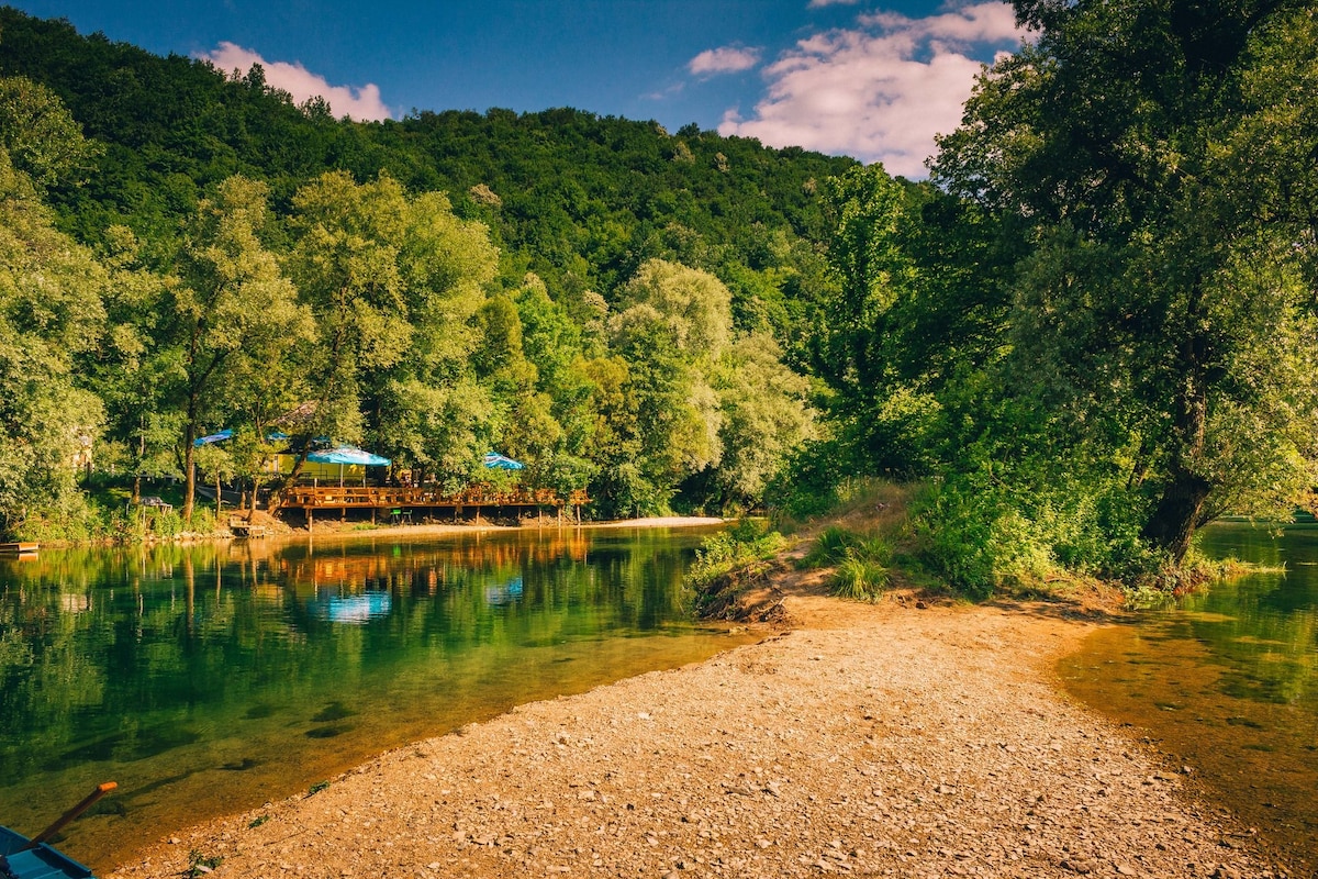 A serene river landscape is captured, showcasing clear waters reflecting surrounding greenery. A beach area of smooth pebbles leads toward the water's edge, with lush trees bordering the riverbank. A wooden deck can be seen in the distance, framed by the rich foliage.