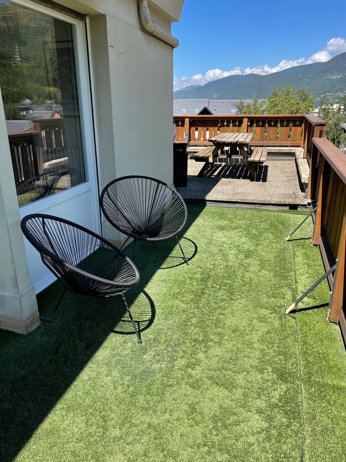 A balcony is featured, showcasing two black wire chairs positioned on artificial grass. A wooden table with several chairs is visible in the background, offering a space for outdoor dining. Scenic mountain views and blue skies enhance the open atmosphere.