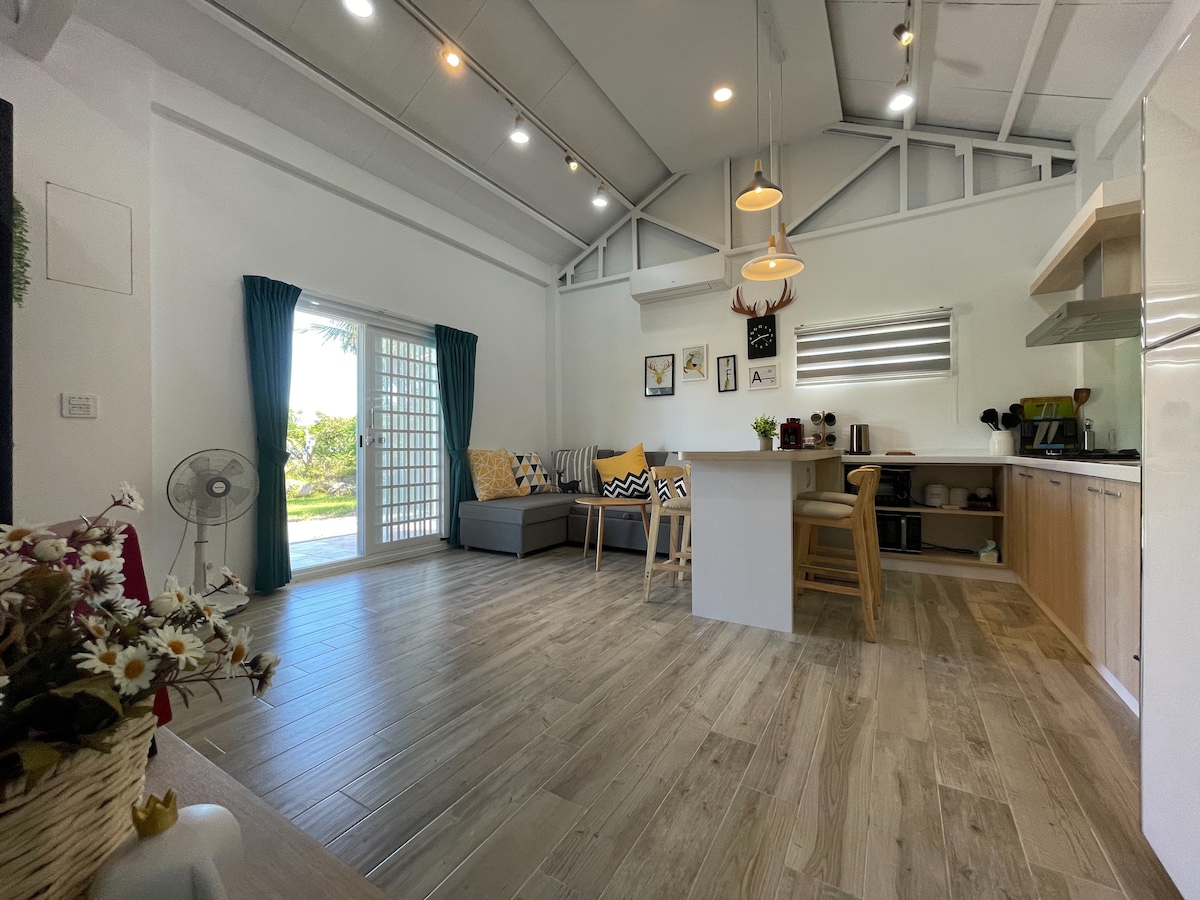 An open living space is highlighted by a comfortable sofa adorned with patterned cushions. Natural light floods in through large glass doors. A dining area features wooden stools at a bar-style counter, while light wooden flooring adds warmth to the atmosphere.