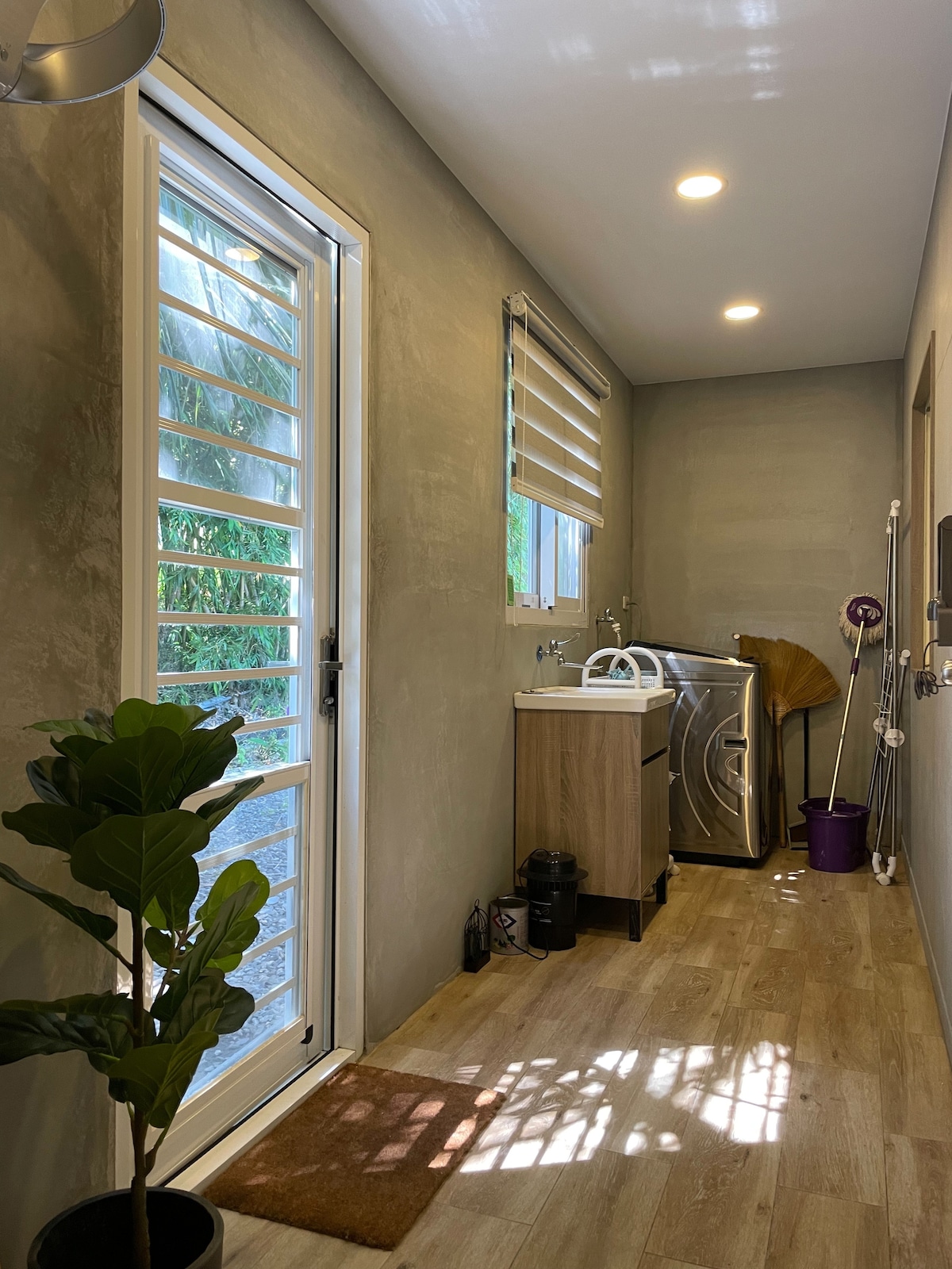 A utility area features a washing machine tucked against a wall, accompanied by a compact wooden storage unit. Natural light enters through a glass door with horizontal slats. A small potted plant and a purple cleaning bucket add touches of greenery and color to the space.