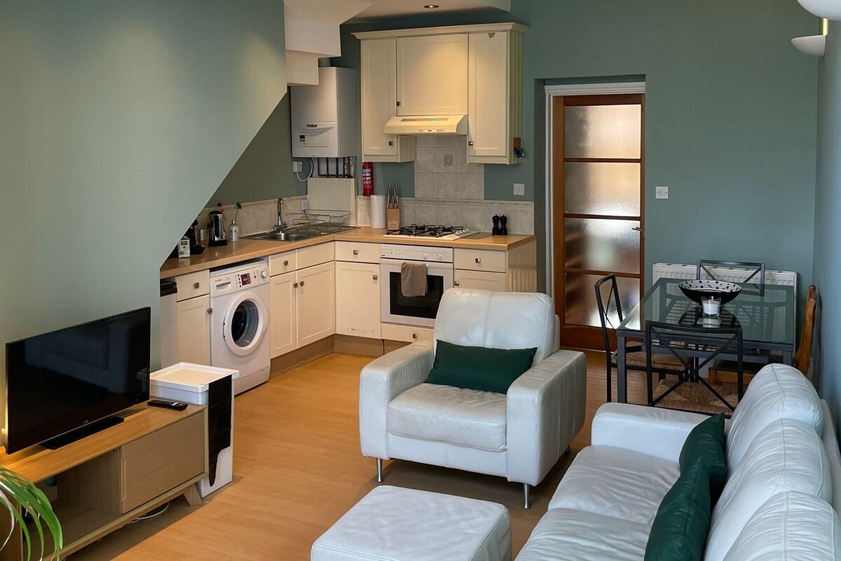 An open-plan living area is featured, showcasing a modern kitchen with white cabinetry and stainless-steel appliances. A comfortable white leather sofa is arranged around a sleek coffee table, and a glass dining table is set nearby. Light-colored flooring completes the inviting space.