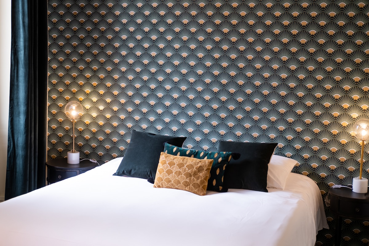 The room features a spacious bed adorned with contrasting pillows, set against a geometric patterned wall. Two stylish bedside lamps provide soft lighting, complemented by dark curtains that frame the window, creating a cozy and inviting setting.