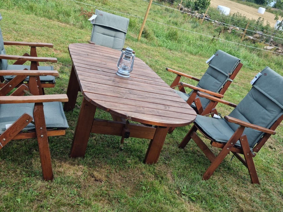 An outdoor dining set is arranged on a grassy area, featuring a wooden table surrounded by six folding chairs. A lantern is placed centrally on the table, enhancing the rustic ambiance, with fields and greenery visible in the background.