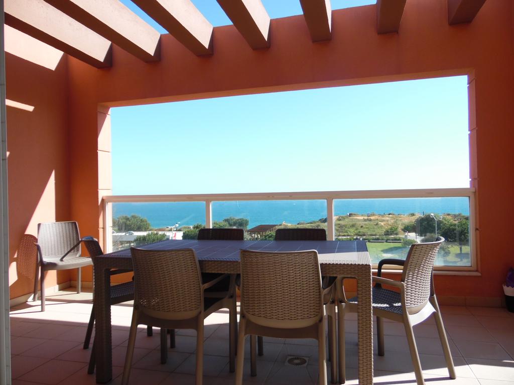 A spacious outdoor terrace features a large table surrounded by several chairs, providing panoramic views of the ocean and landscape. The scene is enhanced by natural light streaming in through wide windows, and architectural elements include wooden beams overhead that offer a sense of openness.