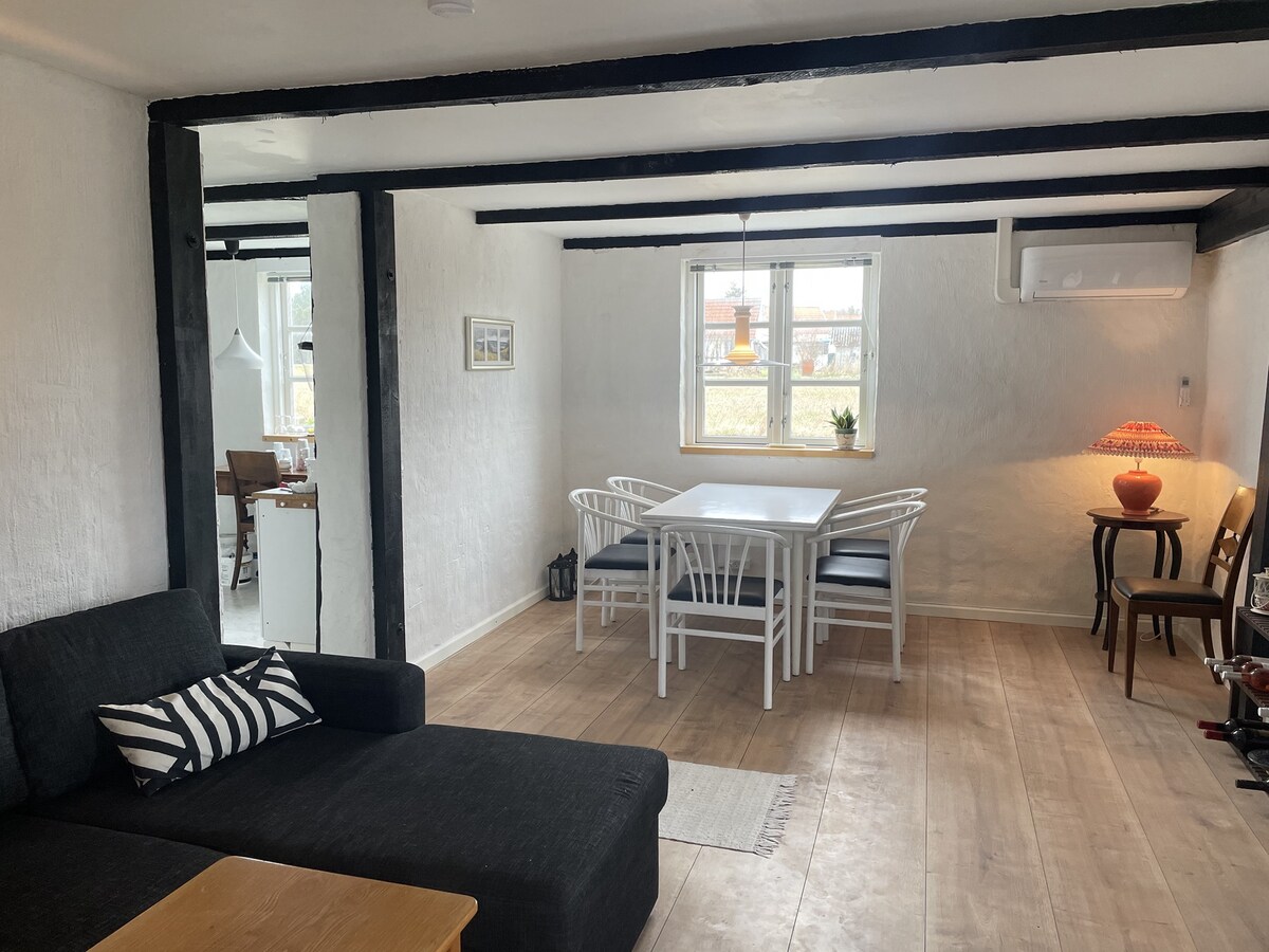 A bright living area is captured, featuring a dark sofa and a stylish coffee table. A white dining table surrounded by chairs is positioned near a window, with greenery visible outside. Exposed beams add character to the ceiling, and a lamp provides soft lighting.