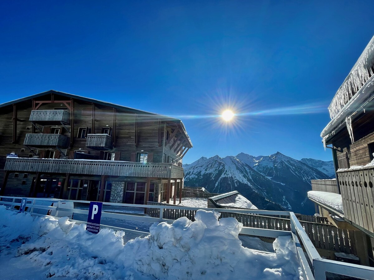 The exterior of a chalet is shown under a clear blue sky, with the sun positioned above the mountains. Snow covers the ground, and wooden balconies provide a rustic aesthetic. A parking sign is visible in the foreground, contributing to the accessible atmosphere.