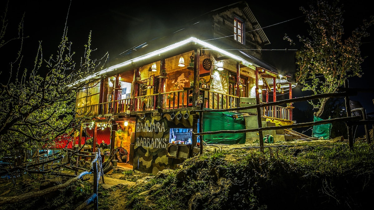 The traditional wood and stone house is illuminated by warm lights, creating a welcoming ambiance at night. The multi-level structure features a wide porch with wooden railings and is surrounded by lush greenery, while apple trees add to the serene atmosphere.