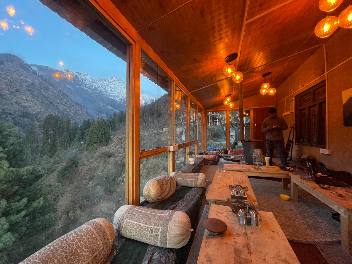 A sunlit room features large windows offering panoramic views of the surrounding mountains. Wooden tables are arranged with cushions for seating. Soft lighting from pendant lamps enhances the cozy atmosphere. Natural elements are incorporated throughout the space, blending with the outdoor scenery.