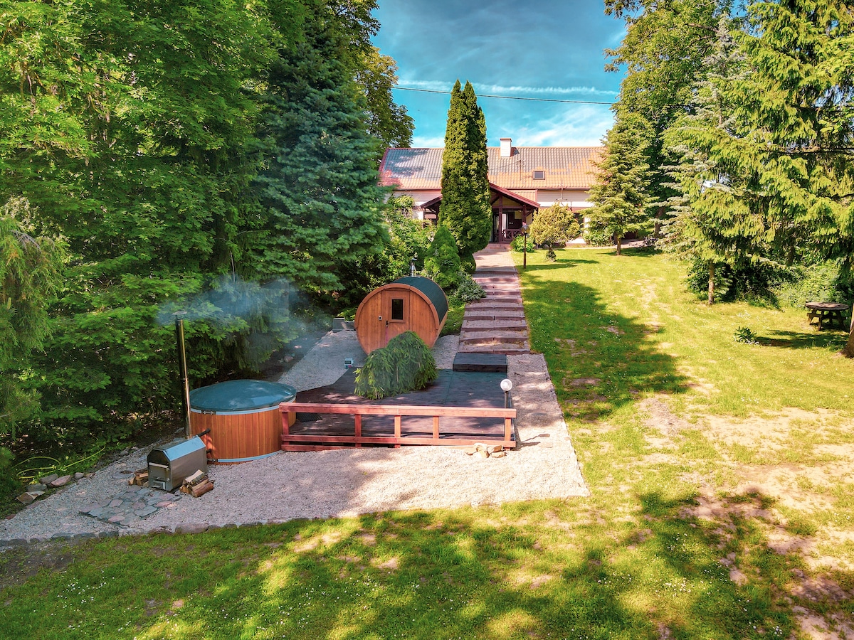 An outdoor relaxation area is featured, presenting wooden hot tubs surrounded by lush greenery. A pathway leads to a welcoming building in the background, partially shaded by tall trees. Sunlight filters through the leaves, creating a serene environment.