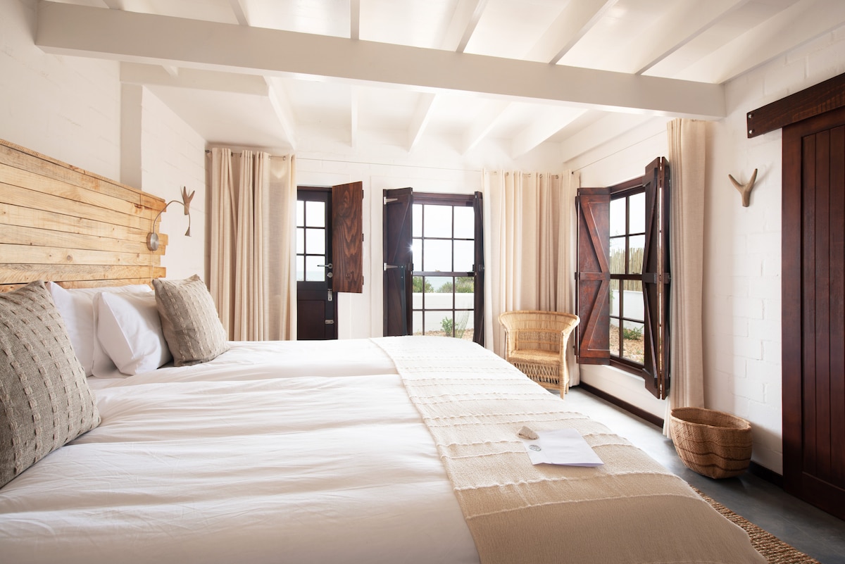 A spacious guest room features two European size beds with neutral bedding. Natural light enters through multiple windows, framed by elegant curtains. Additional elements include a woven chair, wooden accents, and a rustic basket, creating a calming environment with views of the surrounding landscape.
