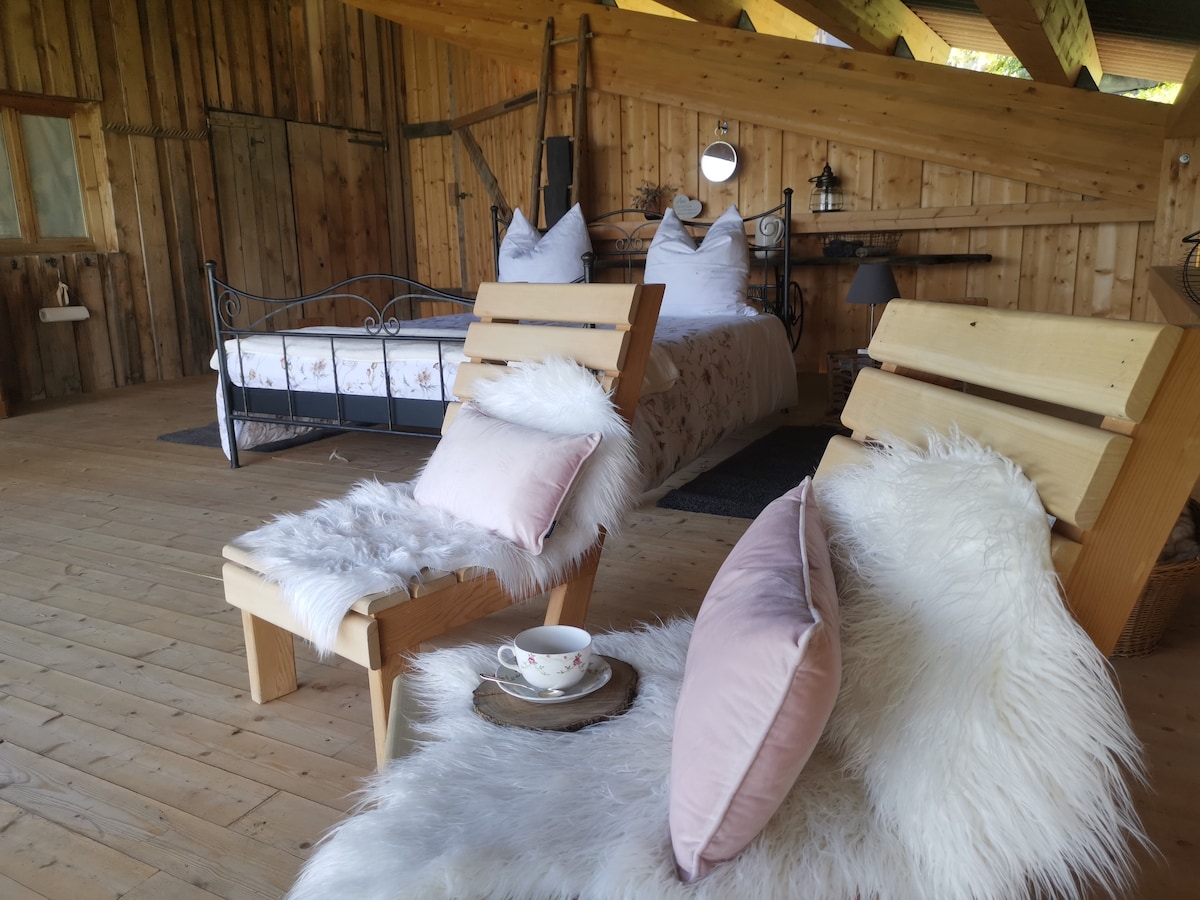 A charming room features a cozy double bed with plush pillows and a warm duvet. Two wooden lounge chairs are placed on a light wooden floor, accented by soft, white throws and pink cushions. Natural light enters through a window, enhancing the rustic ambiance.