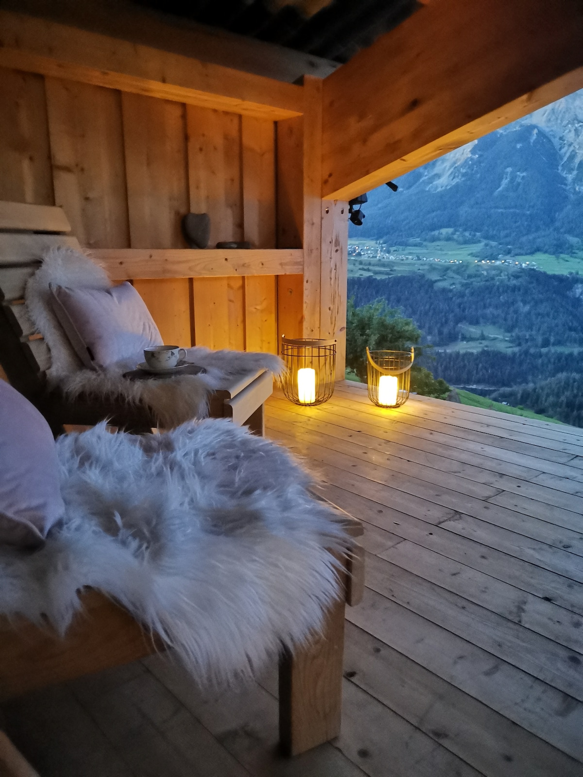 A wooden balcony is featured, adorned with two cozy chairs draped in soft throws. Warm lanterns provide gentle illumination, while a scenic view of the mountains and valley is visible in the background, enhancing the calming atmosphere of the space.