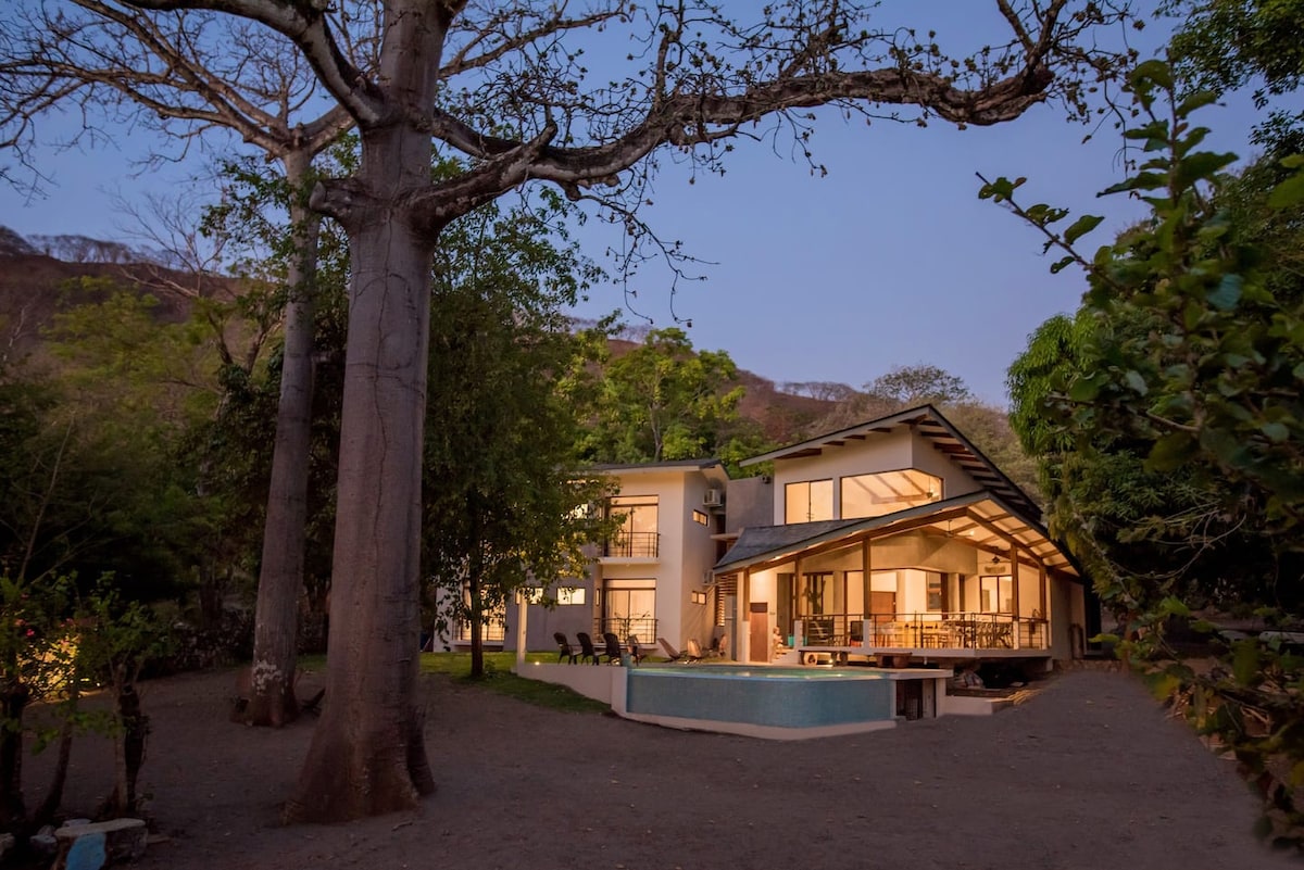 The modern two-story villa is set within a spacious garden, surrounded by trees. Large windows reflect warm light from within. A private swimming pool is visible in the backyard, complemented by an outdoor dining area and a cozy fire pit, enhancing outdoor leisure options.