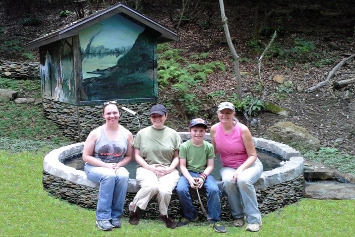 A circular stone seating area is situated on a grassy patch. In the background, a small structure featuring mural artwork is visible among lush greenery. Four individuals are seated together, creating a relaxed atmosphere within the natural setting.