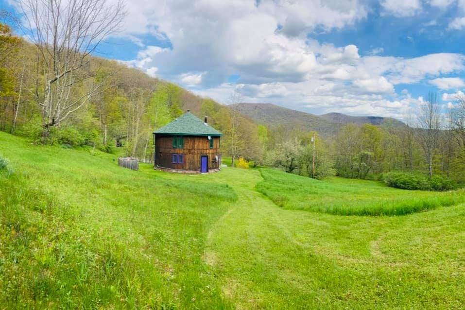 A circular cabin with a green roof is set amidst a lush landscape of rolling green hills and trees. The cabin features a blue door and is surrounded by open fields, with gentle slopes leading to distant mountain ranges under a partly cloudy sky.